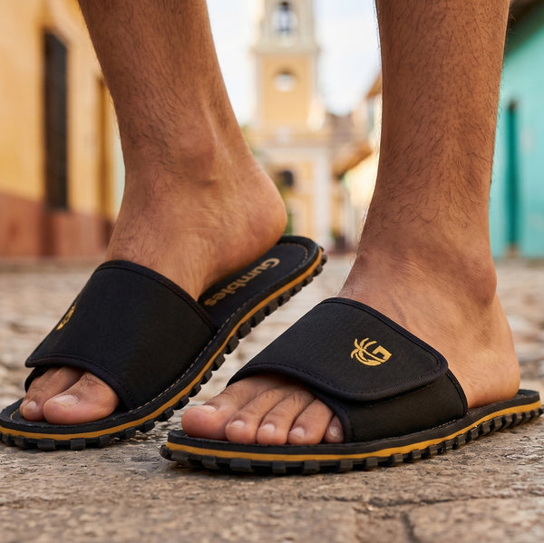 GUMBIES FRANCE Strider black slides for men, featuring a black canvas upper with adjustable strap and gold embroidered palm tree logo. Comfortable EVA sole and recycled rubber outsole.