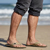 GUMBIES FRANCE tan canvas flip-flops with black soles on a man's feet at the beach.